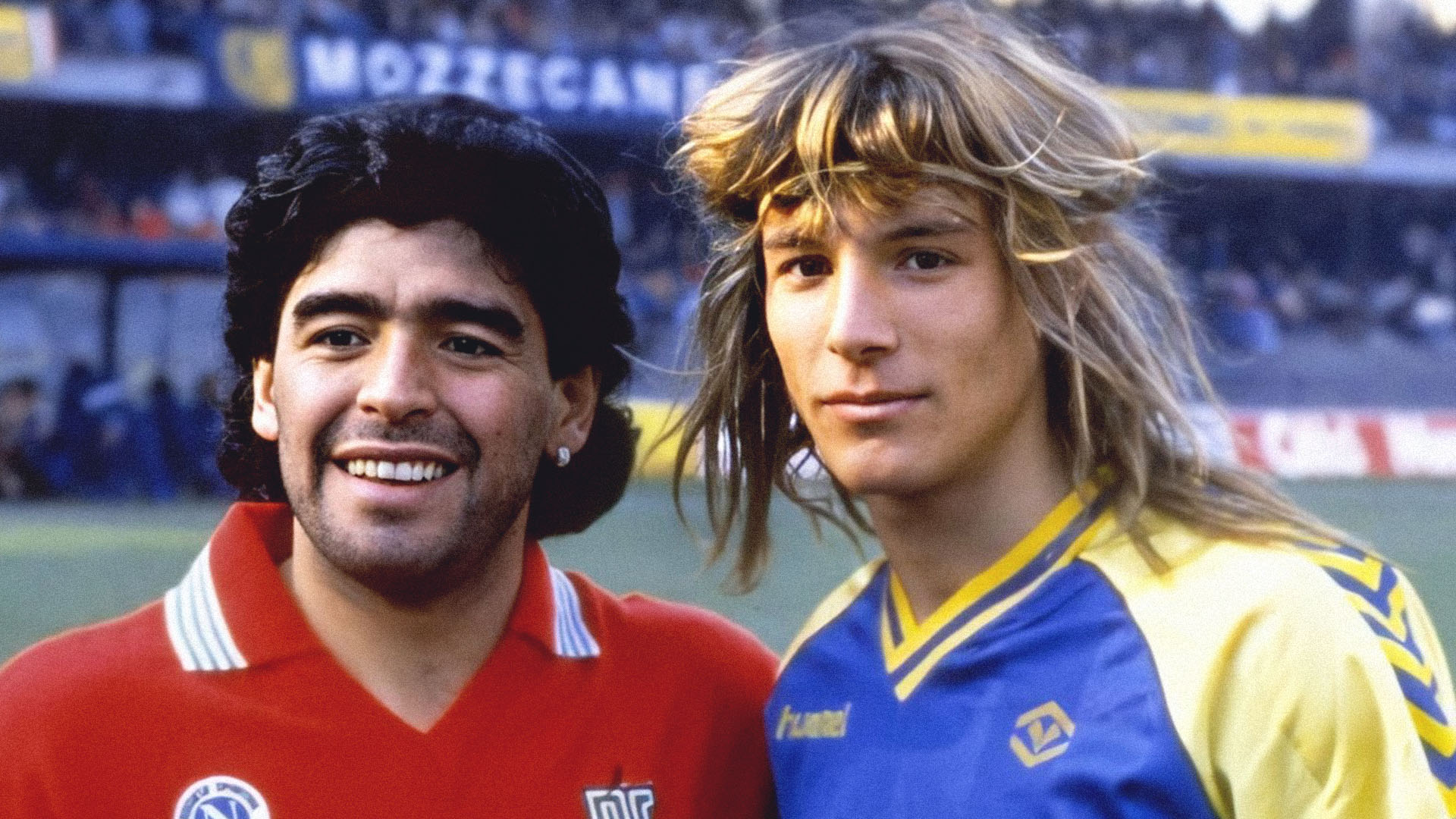 Dois jogadores lado a lado num estádio: à esquerda um de camisola vermelha e cabelo escuro e encaracolado, à direita um jovem de cabelo comprido louro e camisola azul e amarela